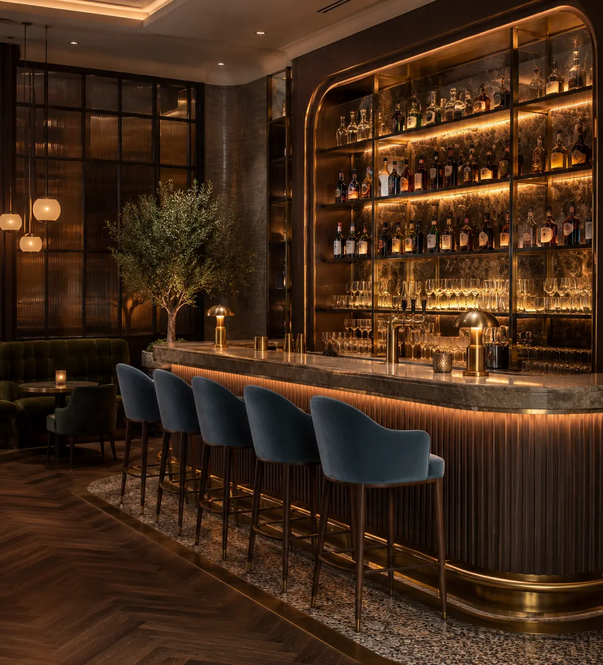 Luxury hotel bar counter with curved marble top and fluted wood front, lined with blue velvet bar stools with brass footrests, backlit glass liquor shelving, brass pendant lights and olive tree accent in an elegant dark wood interior.